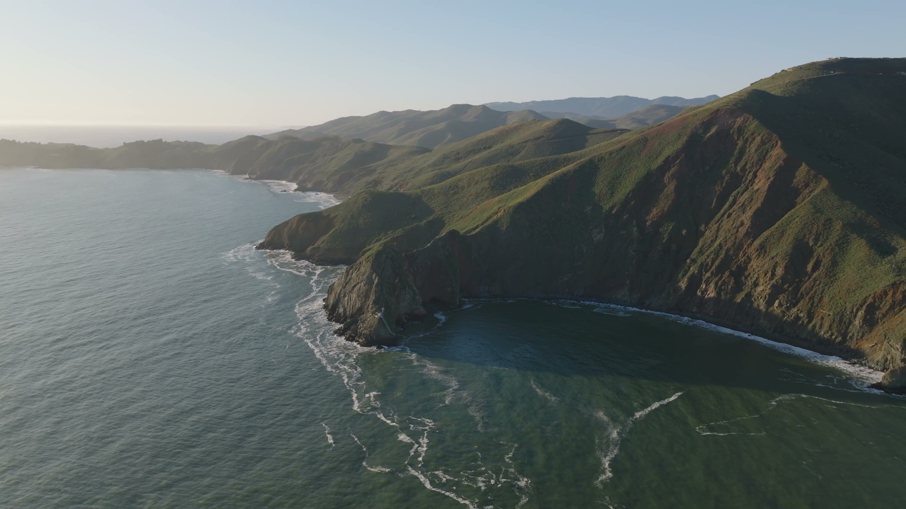 Ocean waves below the grassy Marin Headlands, with the coastline fading into warm coastal haze.